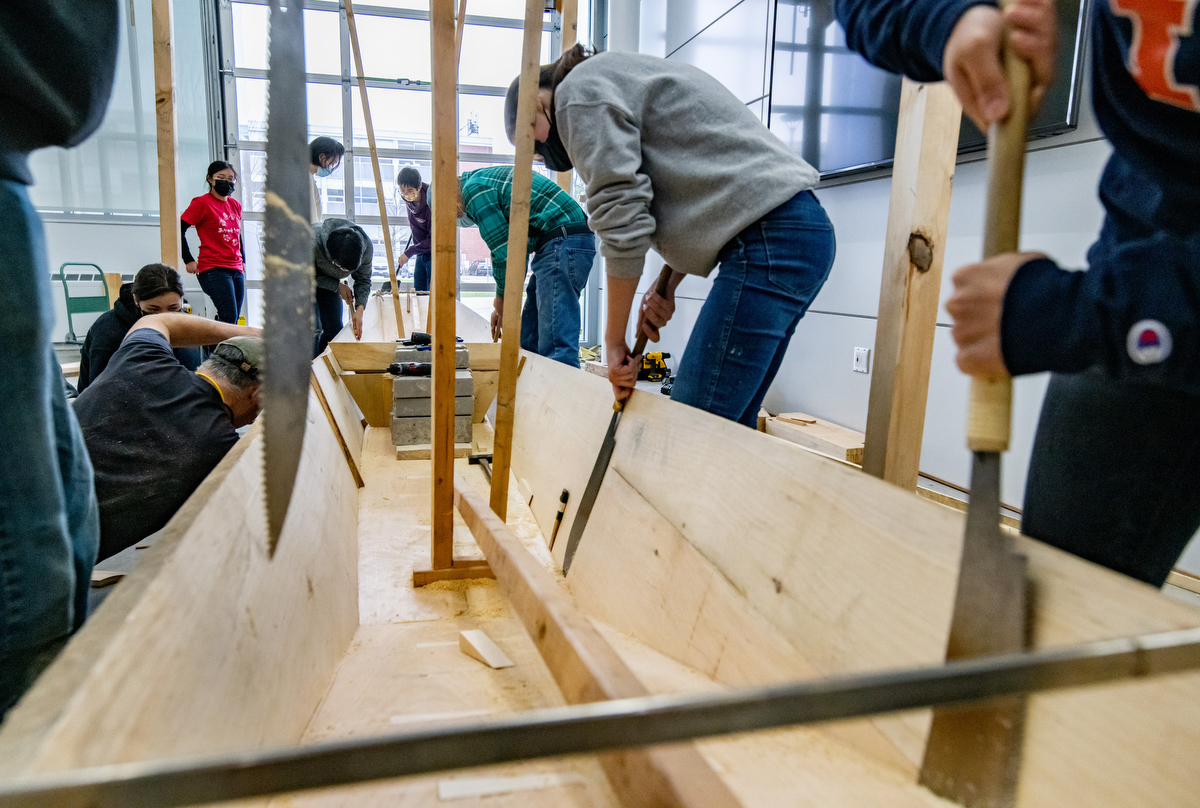Photo of students working along the side of a boat, using hand saws on the seam between the boat’s side and bottom.