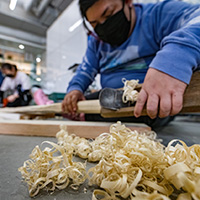 Photo with a pile of wood shavings in the foreground and a student using a hand plane on a piece of wood.