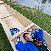 Photo of a student lying in a wooden boat and signing the underside of a beam.