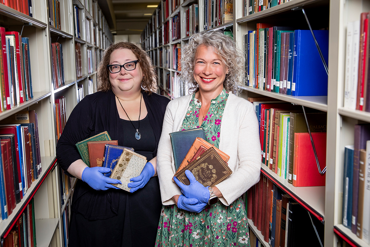 Photo of Shelby Strommer and Jennifer Hain Teper standing between library stacks and each holding several books.