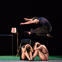 Photo of a male dancer dressed in black leaping over two females sitting on the stage with their hands covering their heads.