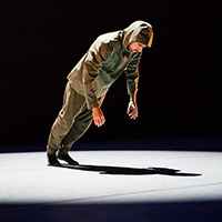Photo of a man in army green pants and a raincoat with the hood up leaning at a 45-degree angle toward the stage floor and casting a shadow beneath him.
