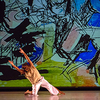 Photo of a stage with abstract art projected onto the back wall, and with a male dancer to the left on one knee, with one leg outstretched to the side and his arms stretched above his head to the other side. Three female dancers to the right have their arms above their heads and are leaning toward center stage.