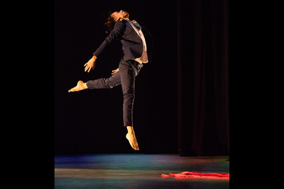 Photo of a male dancer dressed in a black suit coat and pants and a white shirt leaping above the stage with one leg bent behind him, his head thrown back and his arms oustretched behind him. A red scarf lies on the stage floor.