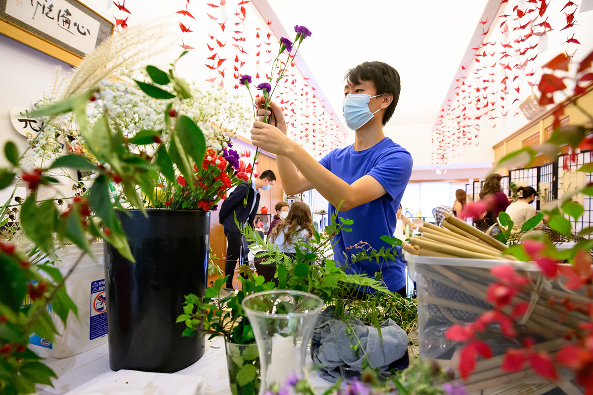 Photo of a student choosing flowers from a table filled with vases of flowers and greenery.  Chains of red paper cranes hang a few feet from the ceiling on both sides.