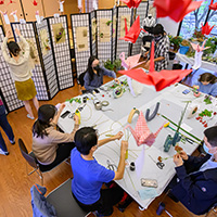 An overhead photo of a table with seated students working on their flower arrangements, while two other students hang their arrangements on a folding screen in the background.