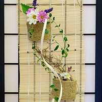 Photo of a hanging flower arrangement tied to a bamboo mat.  Two burlap containers tied to the mat hold flowers, with ivy flowing between the two containers.