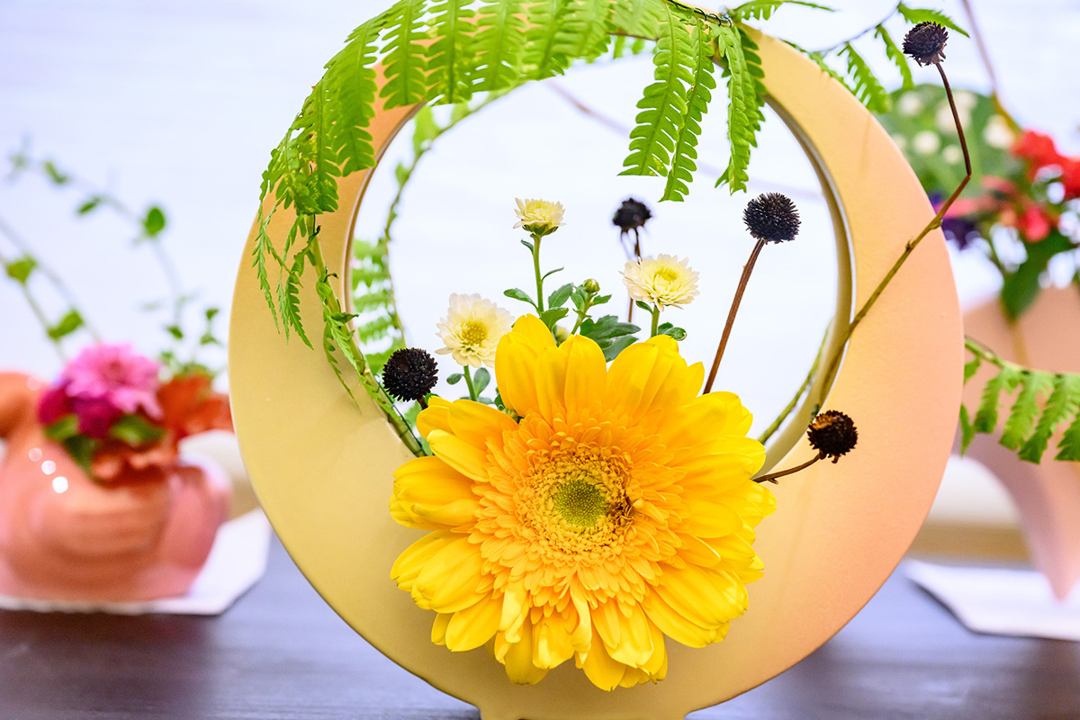Photo of a circular yellow vase containing a large yellow flower and smaller flowers and greenery.