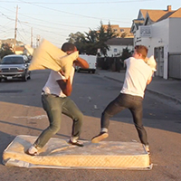 Image of two men standing on a mattress in the middle of a street, having a pillow fight.
